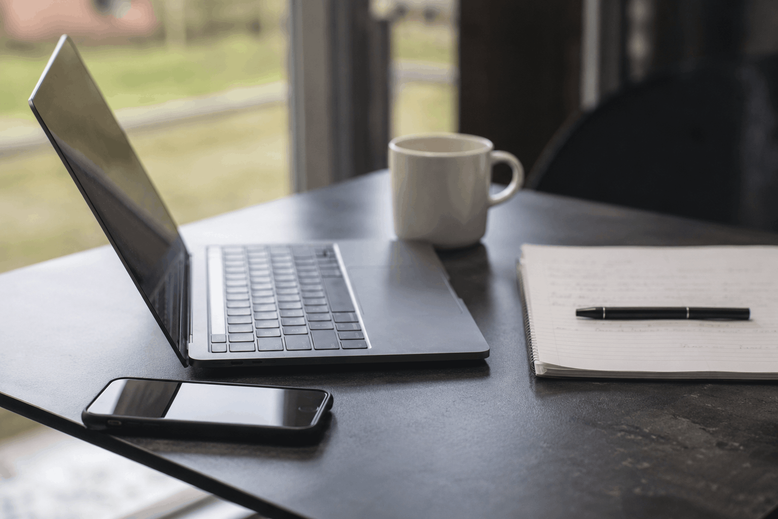 Laptop, mobil, anteckningsblock, penna och kaffekopp på ett bord vid ett fönster